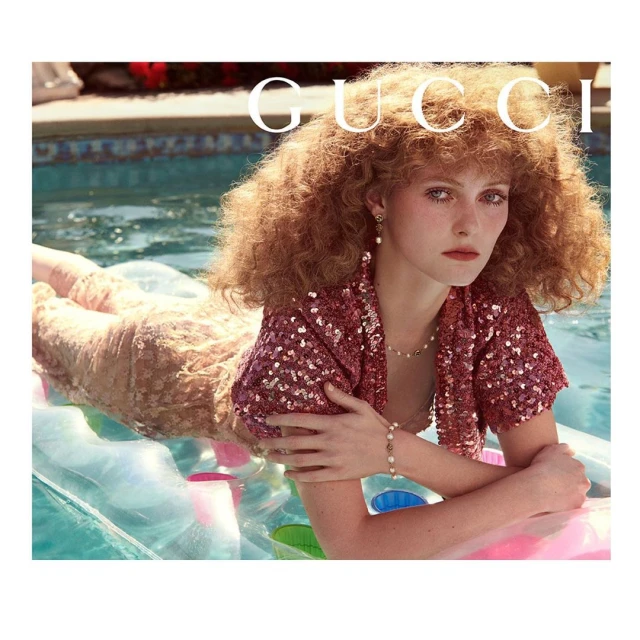 Adult female model with auburn curly hair reclines on an inflatable raft in a swimming pool, wearing a sequined jacket and pearl jewelry.