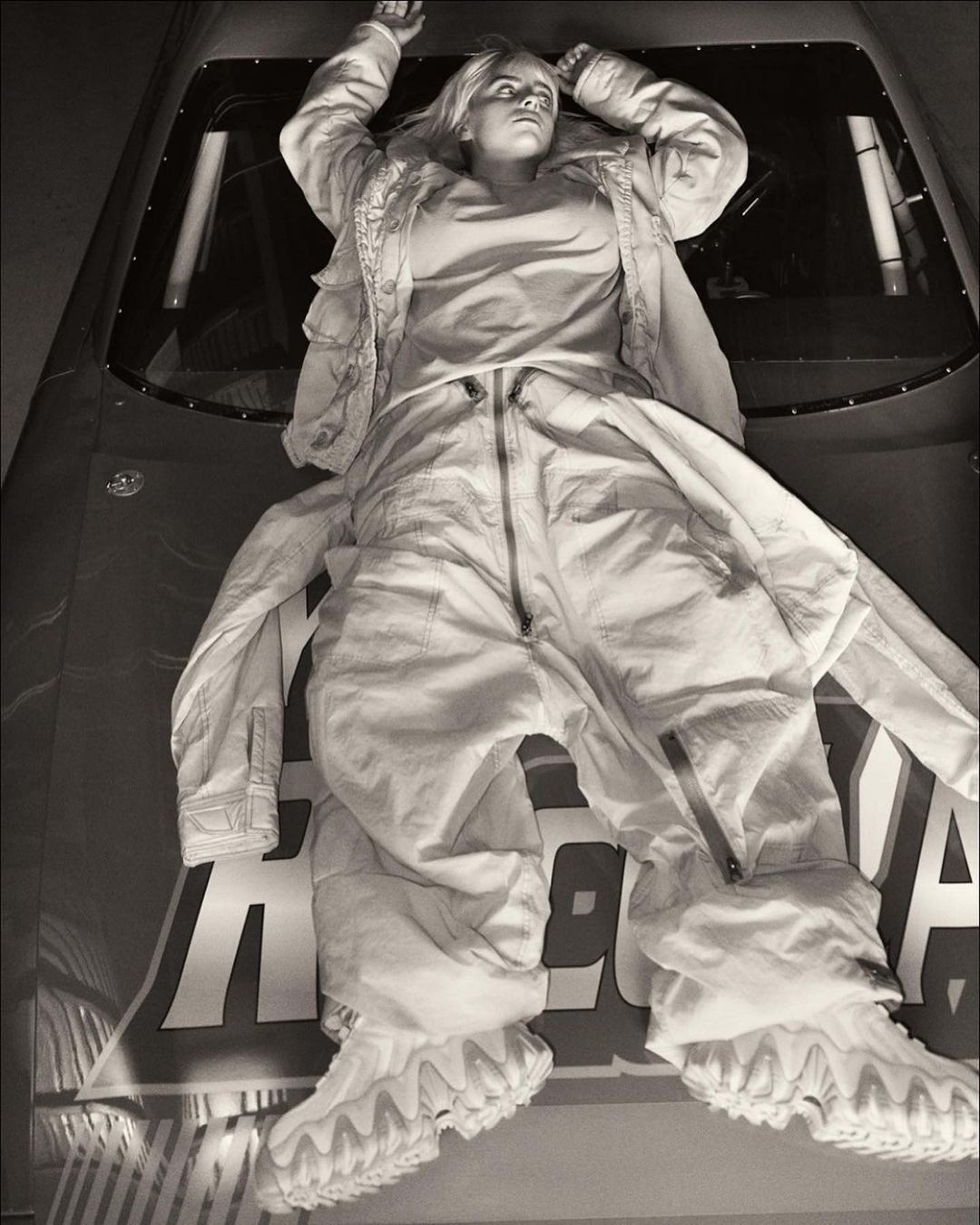 Monochrome studio portrait of a person lying on a car hood in a white jumpsuit and jacket with hands raised, dramatic lighting, and bold text.