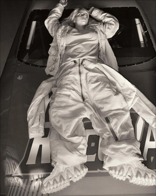 Monochrome studio portrait of a person lying on a car hood in a white jumpsuit and jacket with hands raised, dramatic lighting, and bold text.