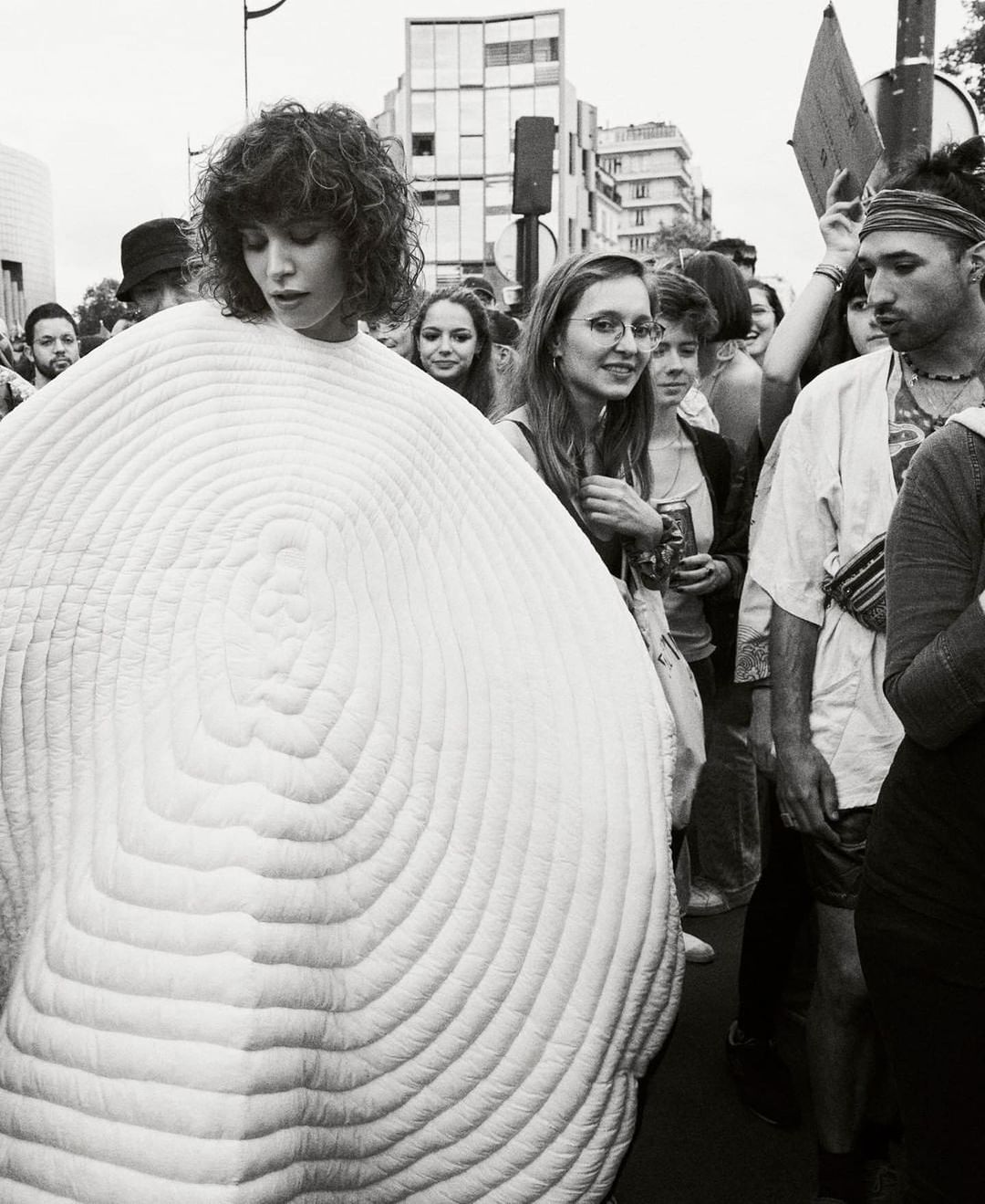 Black and white photo of a woman with curly hair wearing a large, round, white textured garment, surrounded by a crowd in a city.