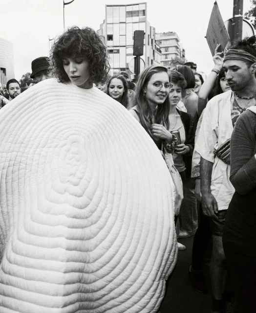 Black and white photo of a woman with curly hair wearing a large, round, white textured garment, surrounded by a crowd in a city.