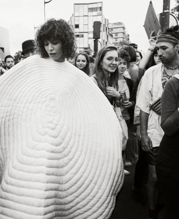 Black and white photo of a woman with curly hair wearing a large, round, white textured garment, surrounded by a crowd in a city.