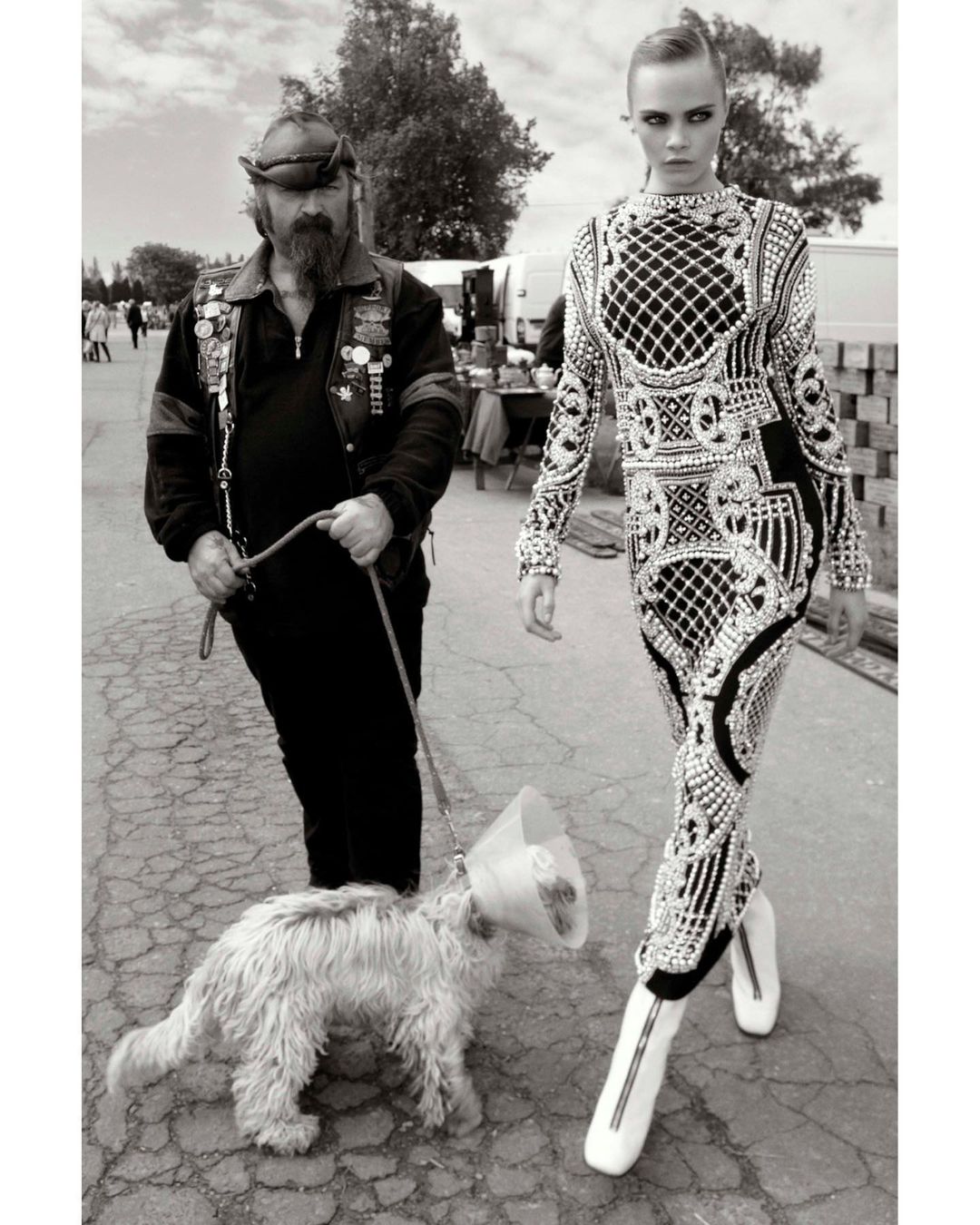 Black-and-white image of a fashion model in an ornate dress walking with a man and a dog on a leash in an outdoor setting.