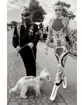 Black-and-white image of a fashion model in an ornate dress walking with a man and a dog on a leash in an outdoor setting.