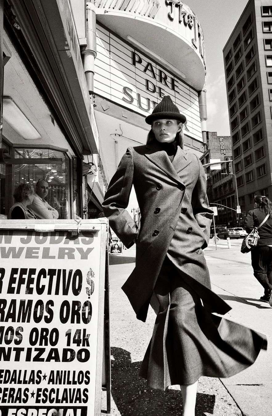Fashionable woman in a coat and hat stands on a city street with a jewellery shop and theatre visible in black and white.