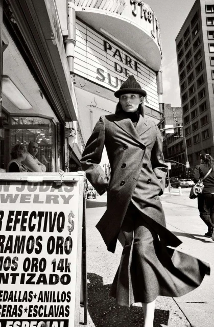 Fashionable woman in a coat and hat stands on a city street with a jewellery shop and theatre visible in black and white.