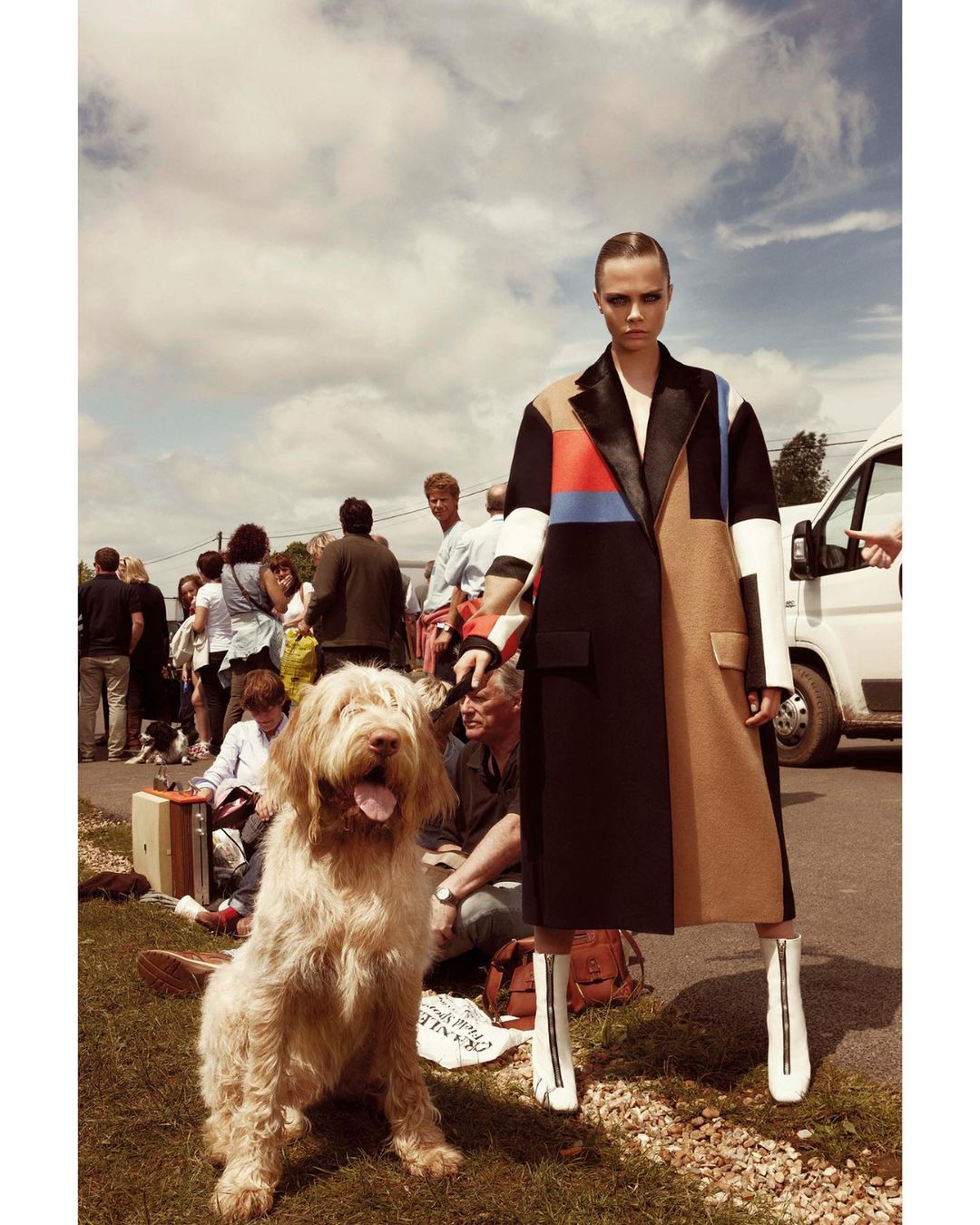 Fashion model poses with a dog in a multicolored coat and white boots on a grassy road. People and a van are in the background.