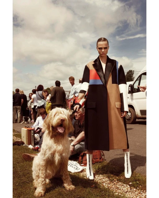 Fashion model poses with a dog in a multicolored coat and white boots on a grassy road. People and a van are in the background.