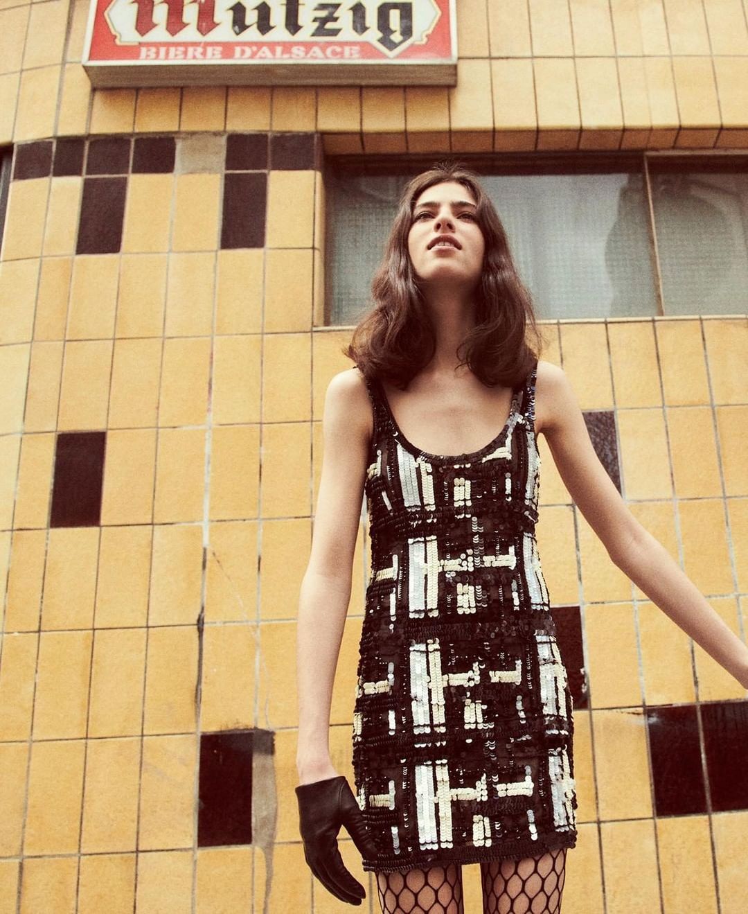 Young woman poses in a sparkly black and white dress against a tiled building. She wears a single leather glove and fishnet stockings.