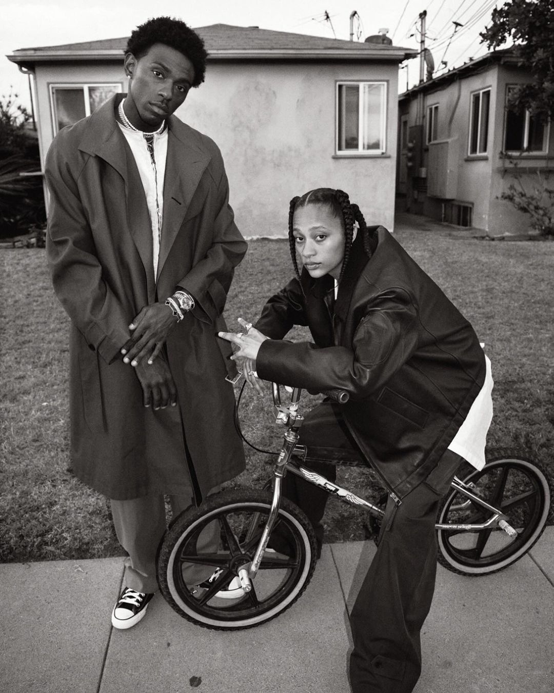 Black and white portrait of two young adults with a BMX bike in an urban setting; the woman poses on the bike, and the man stands next to her.
