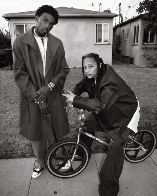 Black and white portrait of two young adults with a BMX bike in an urban setting; the woman poses on the bike, and the man stands next to her.