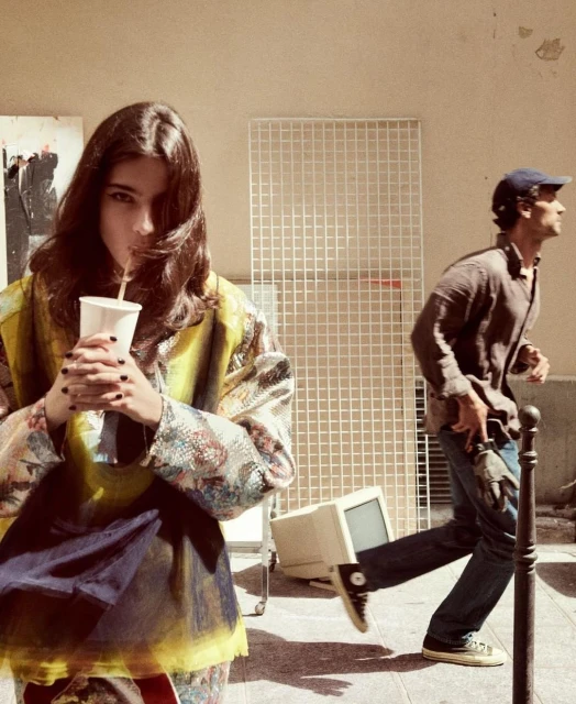 Young woman sips from a paper cup in an urban setting, while a man jogs in the background near a discarded computer monitor.