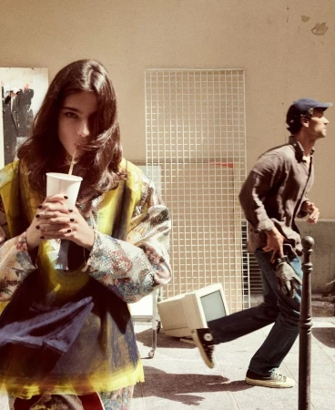 Young woman sips from a paper cup in an urban setting, while a man jogs in the background near a discarded computer monitor.