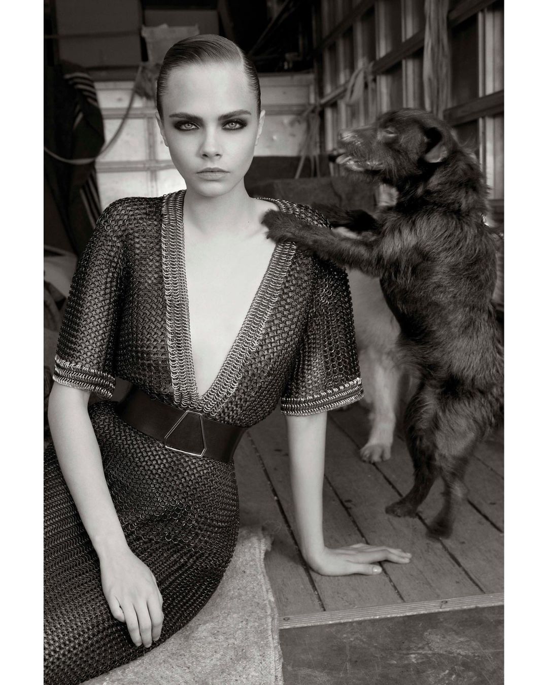 Black and white portrait of Cara Delevingne in a chainmail dress, seated next to a dog that is resting its paw on her shoulder.