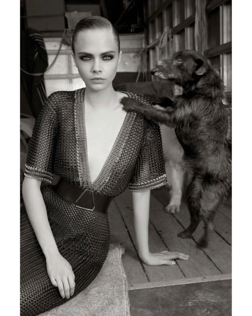 Black and white portrait of Cara Delevingne in a chainmail dress, seated next to a dog that is resting its paw on her shoulder.