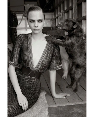 Black and white portrait of Cara Delevingne in a chainmail dress, seated next to a dog that is resting its paw on her shoulder.
