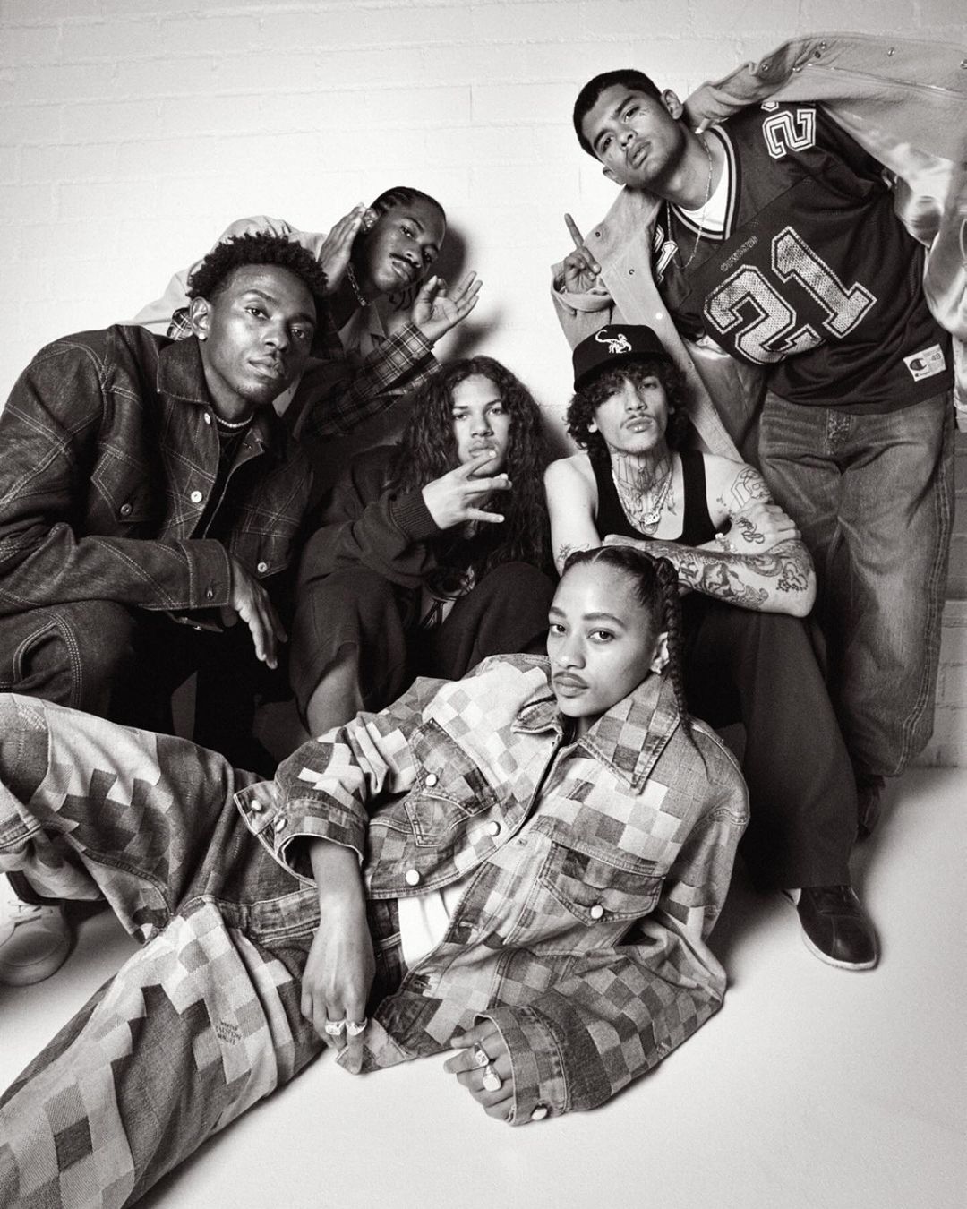 Group shot of seven people posing indoors against a white wall in fashionable denim and streetwear in a black-and-white photograph.