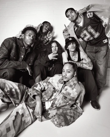 Group shot of seven people posing indoors against a white wall in fashionable denim and streetwear in a black-and-white photograph.