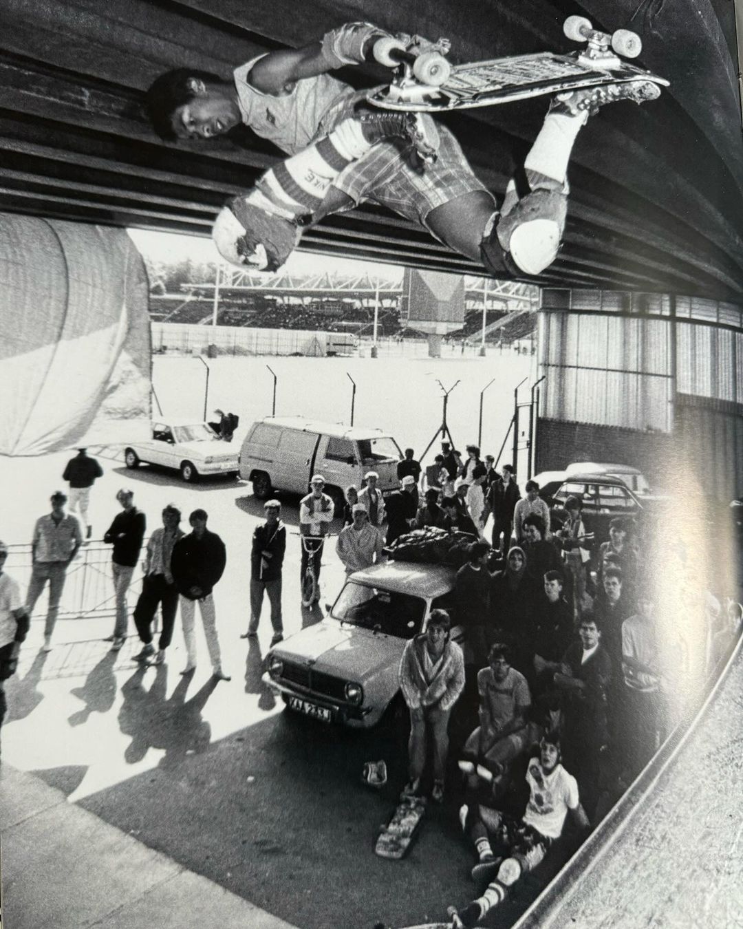 A black and white shot shows a skateboarder in mid-air performing a trick, with a crowd of spectators, cars, and a stadium in the background.