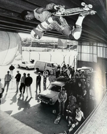 A black and white shot shows a skateboarder in mid-air performing a trick, with a crowd of spectators, cars, and a stadium in the background.