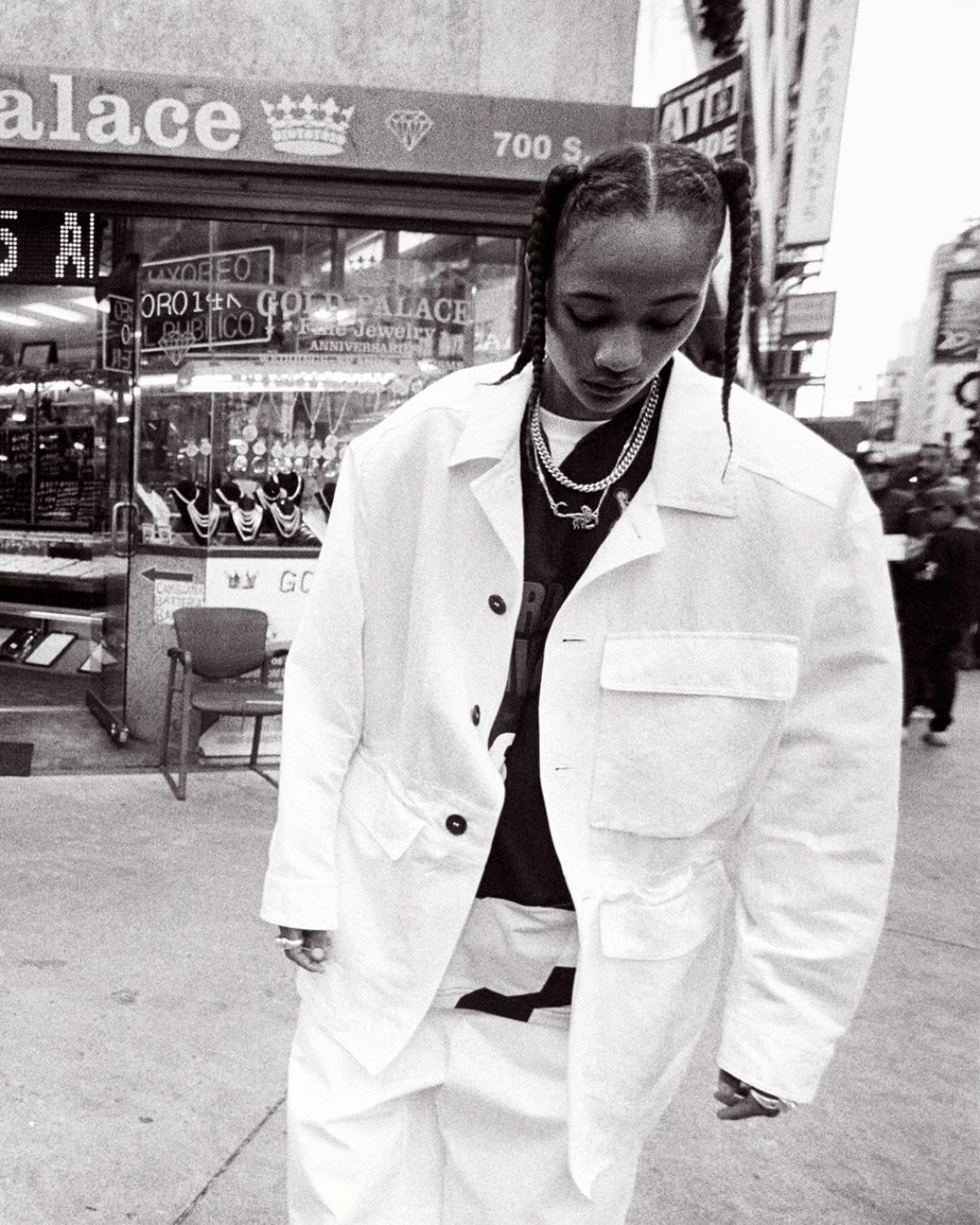 Woman in white jacket and pants stands on city street in front of jewelry store with braid hairstyle and layered necklaces, grayscale image.