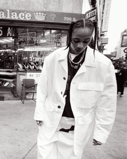 Woman in white jacket and pants stands on city street in front of jewelry store with braid hairstyle and layered necklaces, grayscale image.