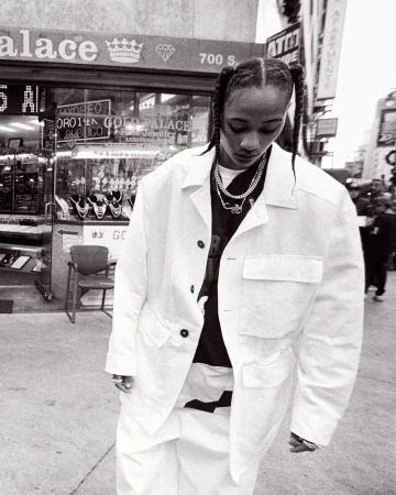 Woman in white jacket and pants stands on city street in front of jewelry store with braid hairstyle and layered necklaces, grayscale image.