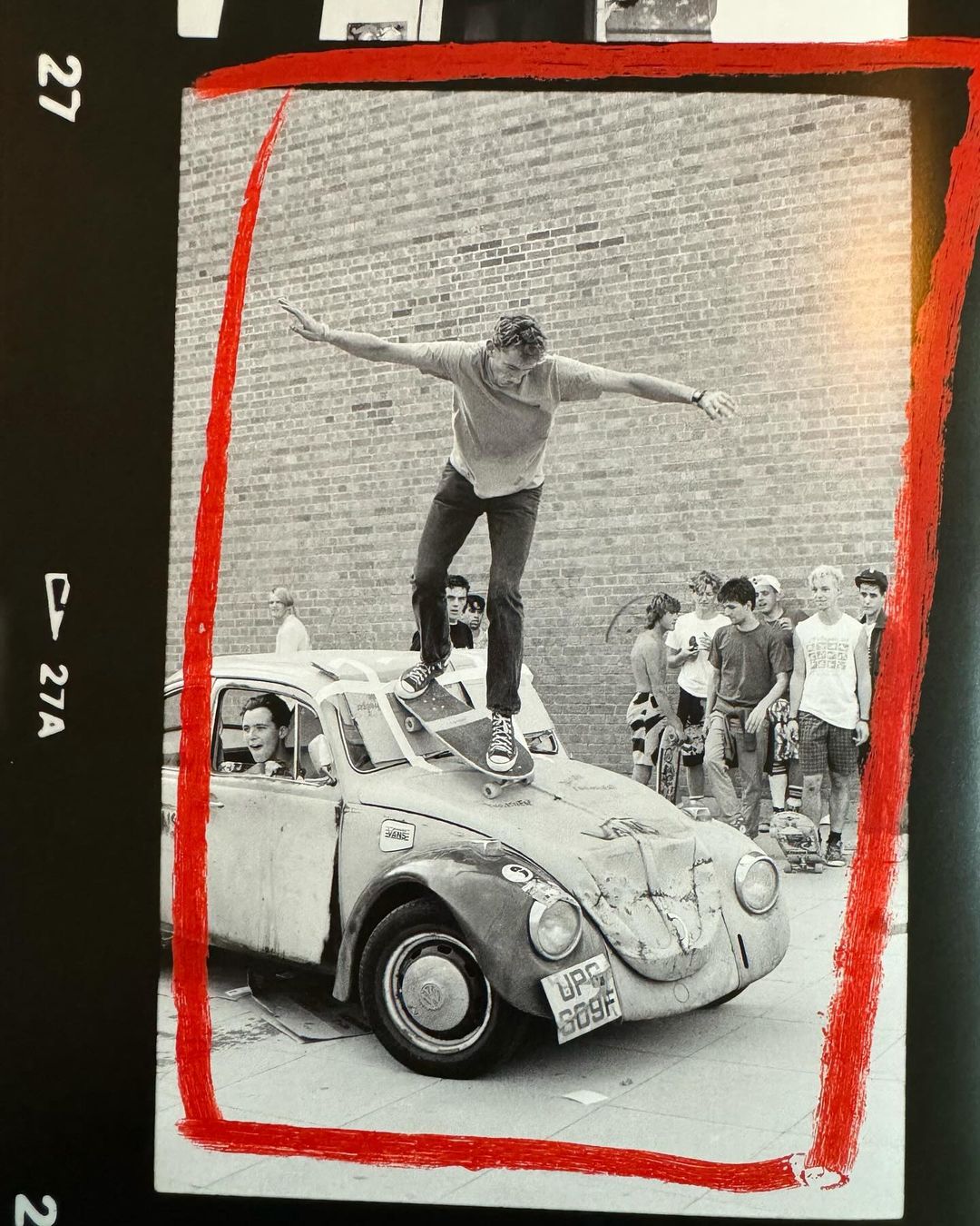 A man stands on a skateboard balanced on top of a vintage car, with other people and a brick wall visible in the background.