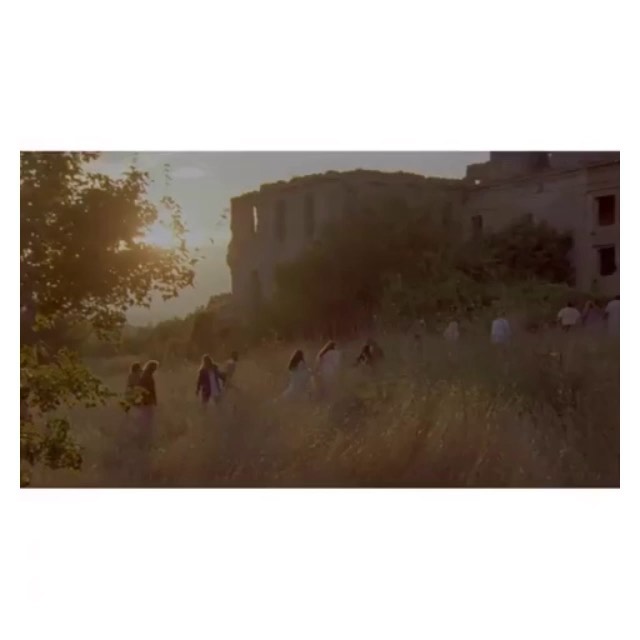 Large group of people walking in a field near an old castle at sunset, bathed in golden light with trees and tall grass.
