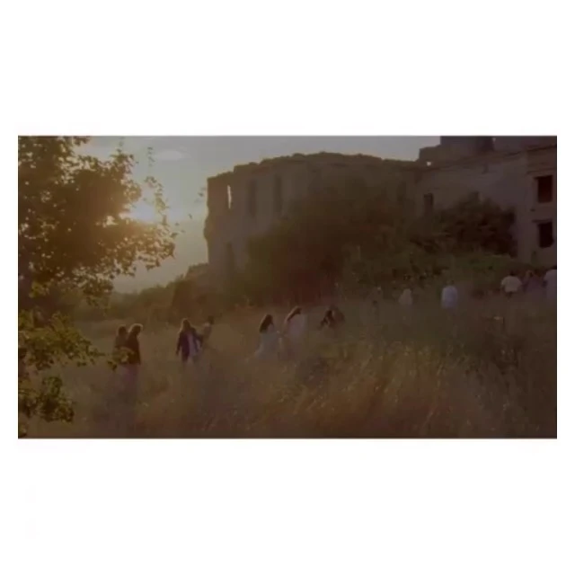 Large group of people walking in a field near an old castle at sunset, bathed in golden light with trees and tall grass.