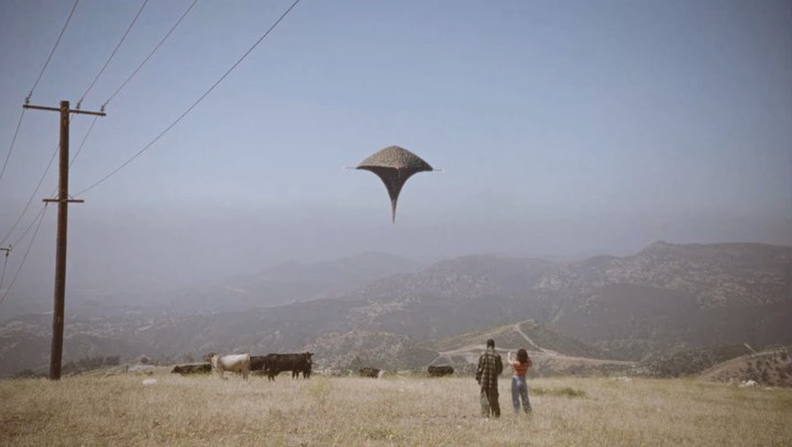 A man and woman stand in a field watching a UFO in the sky above a herd of cows in a rural landscape under a clear blue sky.