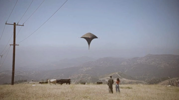A man and woman stand in a field watching a UFO in the sky above a herd of cows in a rural landscape under a clear blue sky.