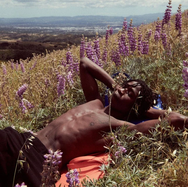An adult man lies in a field of wildflowers on a hillside, enjoying the sunny day with rolling hills and a city in the distance.