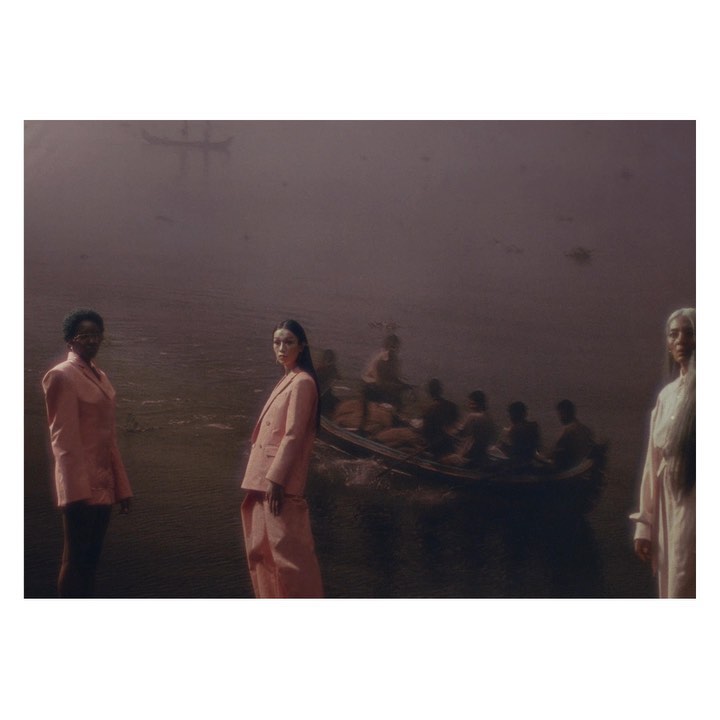Three women in suits pose near water with a boat filled with people in the background, evoking a sense of mystery and contemplation.