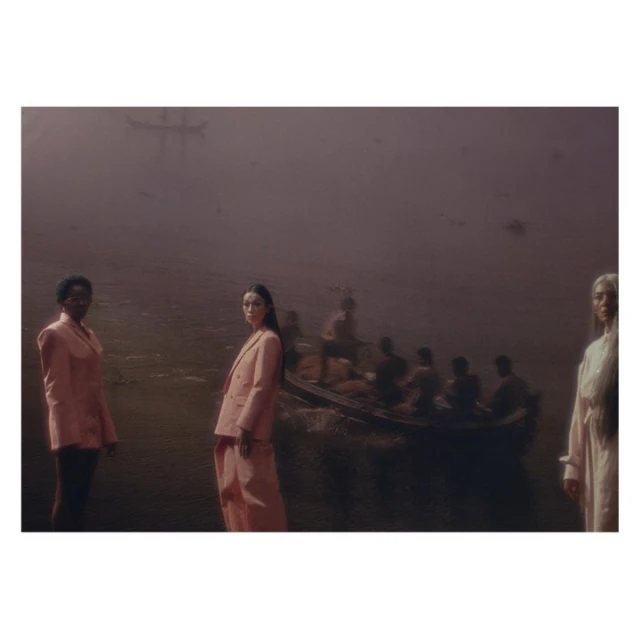 Three women in suits pose near water with a boat filled with people in the background, evoking a sense of mystery and contemplation.