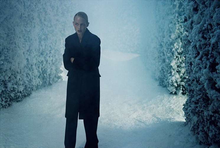 Adult female stands in a snow-covered hedge maze, wearing a dark coat and trousers, with cool, muted tones, creating a serene and wintery image.