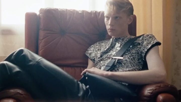 Indoor portrait of a young adult posing in a red leather chair, wearing shiny silver top, and black leather trousers in a dimly lit room.