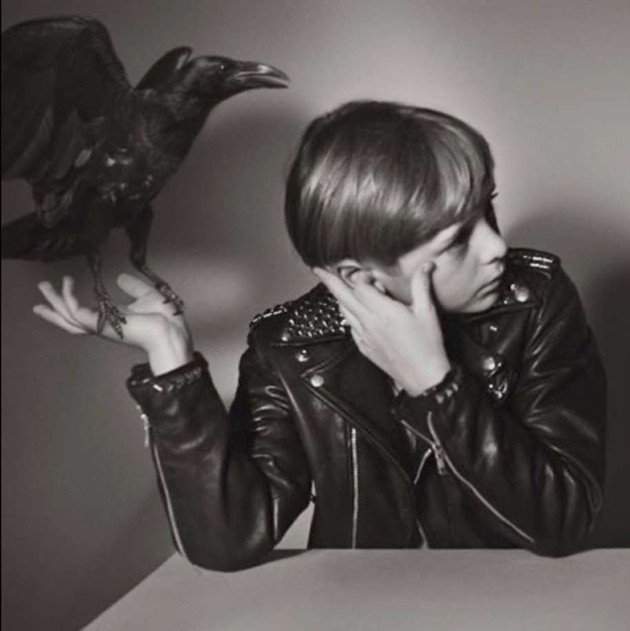 Monochrome shot of person in studded leather jacket with a raven perched on their hand against a neutral background.
