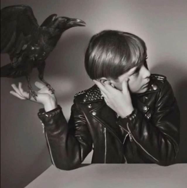 Monochrome shot of person in studded leather jacket with a raven perched on their hand against a neutral background.