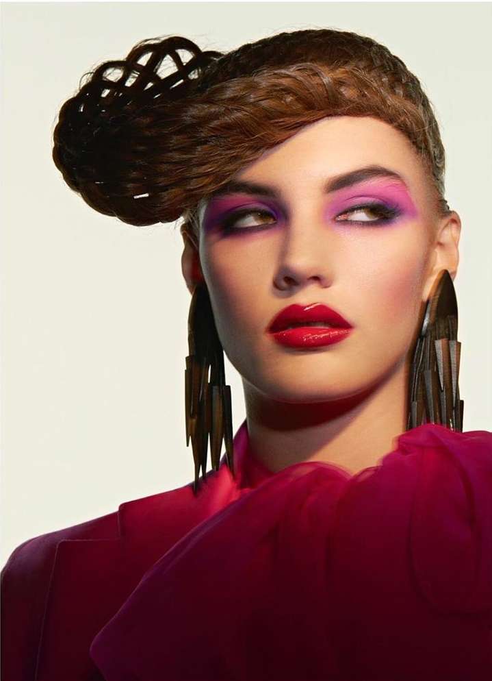 Headshot of an adult female model with creative brown hairstyle, magenta eyeshadow and lipstick, and large geometric earrings.