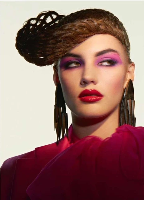 Headshot of an adult female model with creative brown hairstyle, magenta eyeshadow and lipstick, and large geometric earrings.