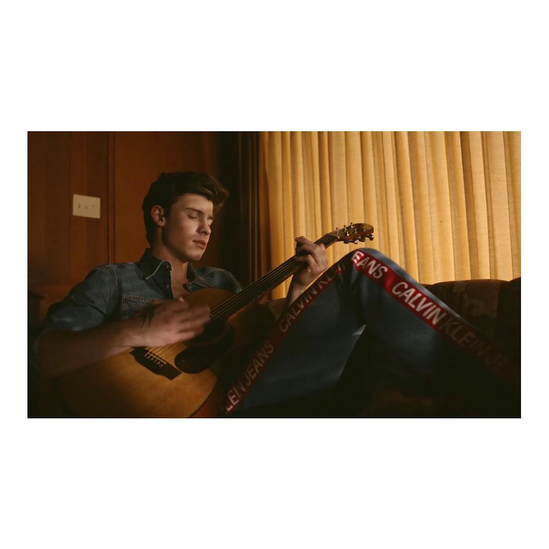 Adult male playing an acoustic guitar while sitting indoors; he wears a denim shirt and jeans branded with the Calvin Klein logo.