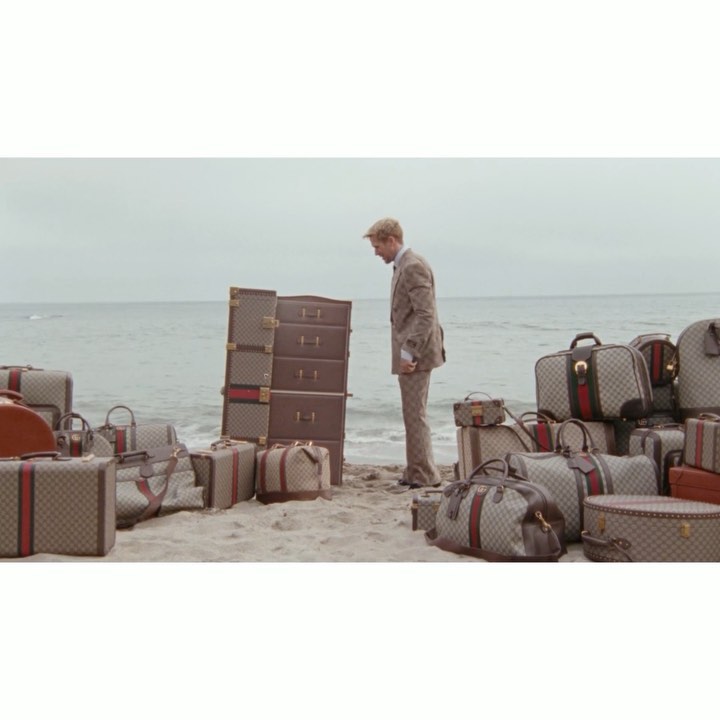 Man in suit stands on a beach by a pile of brown Gucci luggage and bags, the ocean is visible in the background on an overcast day.