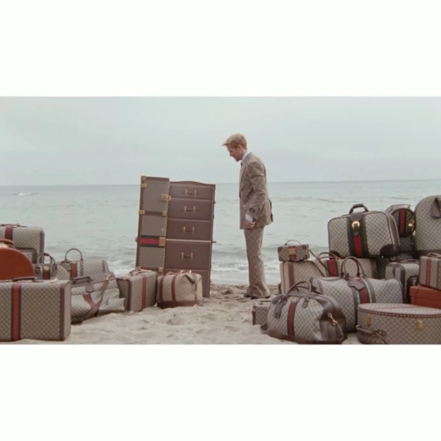 Man in suit stands on a beach by a pile of brown Gucci luggage and bags, the ocean is visible in the background on an overcast day.
