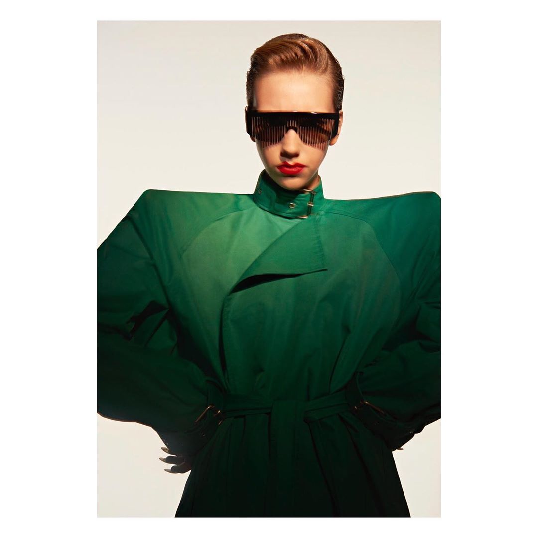 A striking fashion portrait features a young woman with red lipstick and geometric sunglasses, posing in a green trench coat against a white backdrop.
