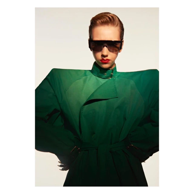 A striking fashion portrait features a young woman with red lipstick and geometric sunglasses, posing in a green trench coat against a white backdrop.