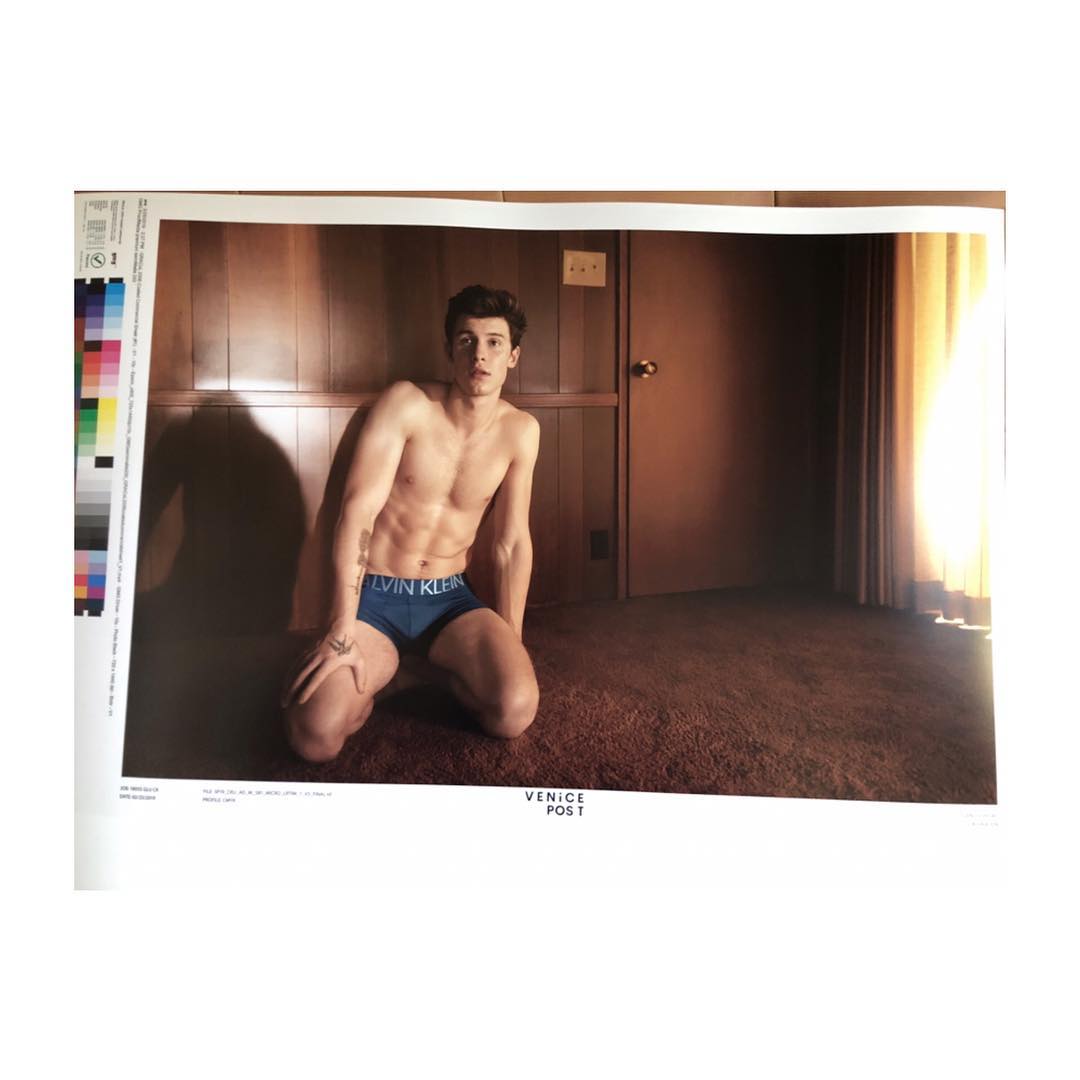 Shirtless man with athletic build kneeling in blue Calvin Klein underwear. Indoor setting, warm color palette, natural lighting.