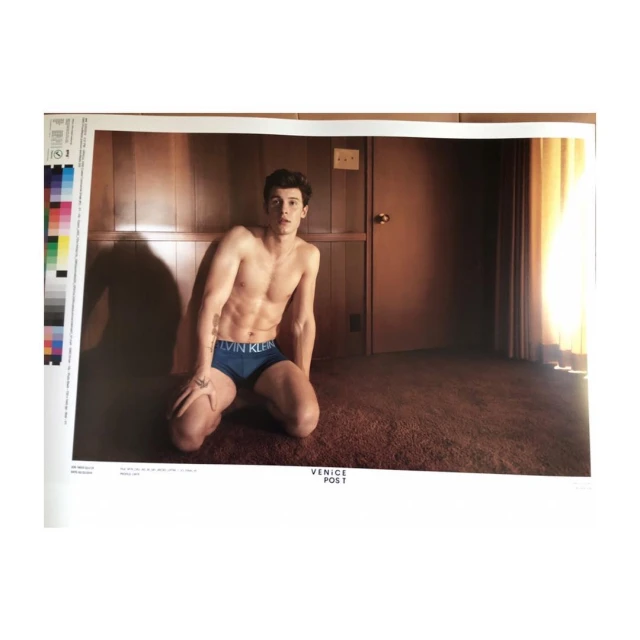 Shirtless man with athletic build kneeling in blue Calvin Klein underwear. Indoor setting, warm color palette, natural lighting.