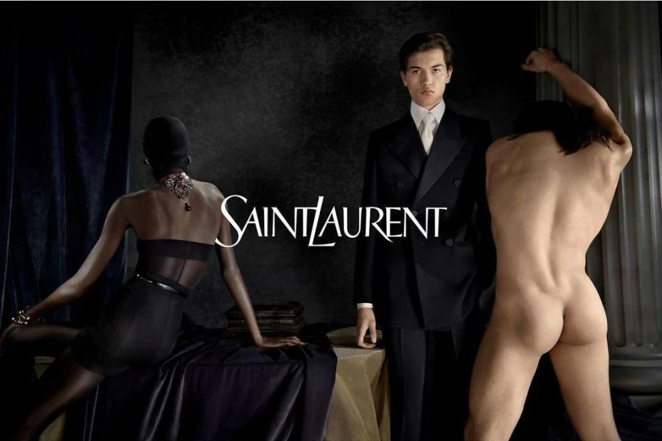 Black and white fashion shot featuring two models flanking a model in a suit against a dark background with the Saint Laurent logo.
