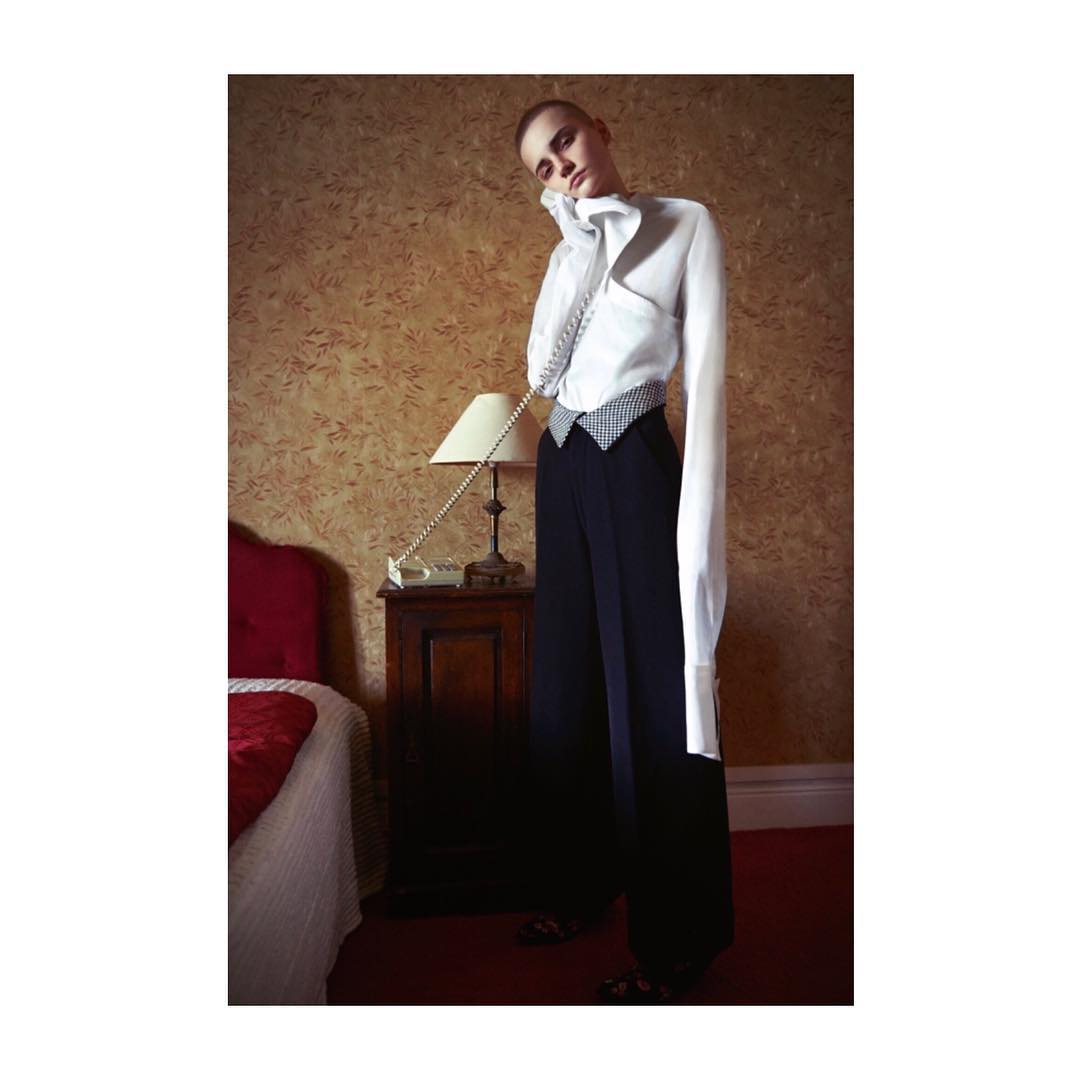 A woman in a white shirt and dark pants poses with a vintage telephone in a room with a bed, creating a fashion editorial image.